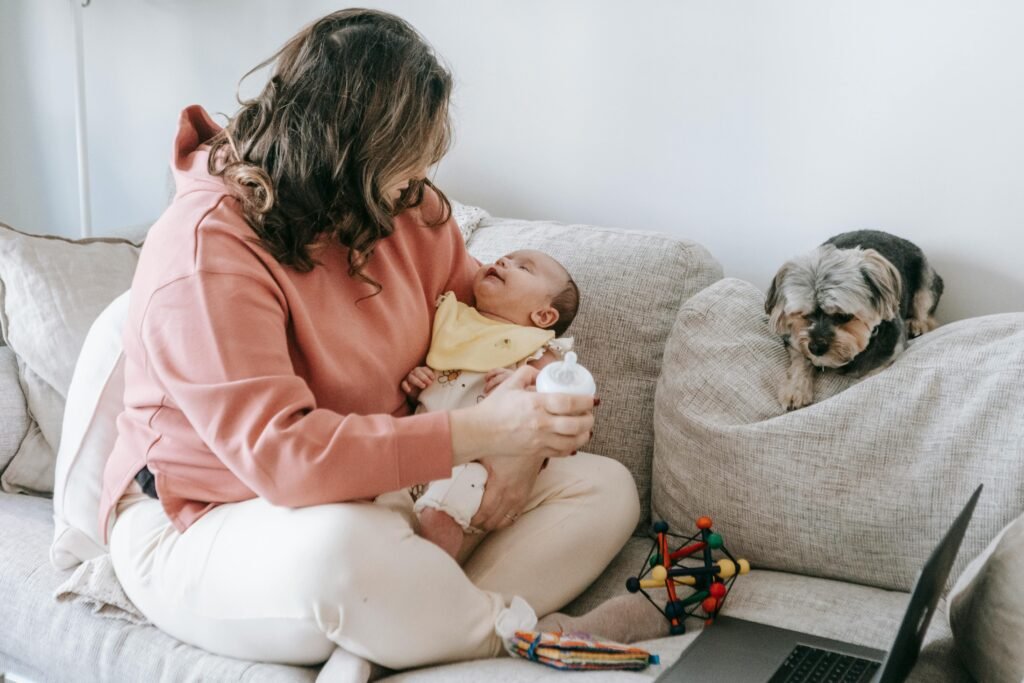 A mother bottle-feeds her baby while sitting on a cozy couch with a small dog nearby.